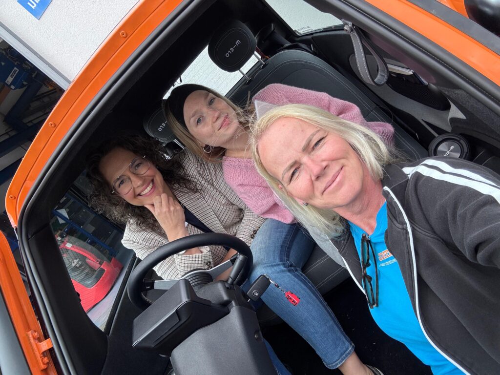 Ein Selfie von drei Frauen in und vor einem kleinen Elektroauto. Der Innenraum ist zum Teil sichtbar.